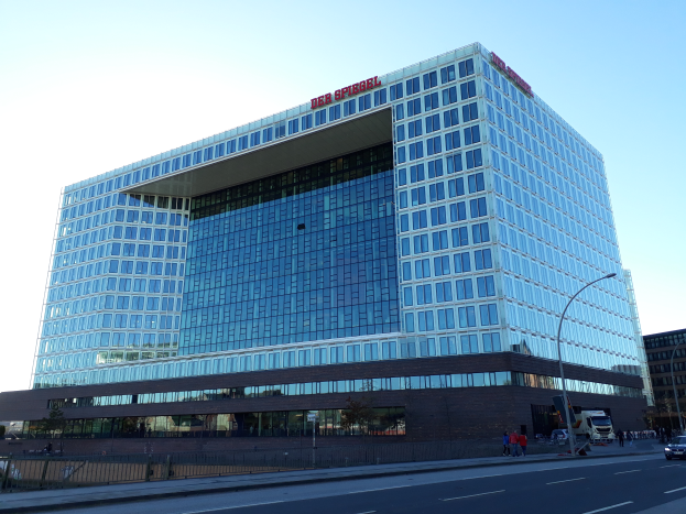 Ein großes Gebäude mit vielen Fenstern, das als Hauptsitz der Deutschen Bank identifiziert wird, umgeben von Straßenlaternen, Straßenlaternen, Kraftfahrzeugen, Fußgängern, Bäumen und einem bewölkten Himmel.