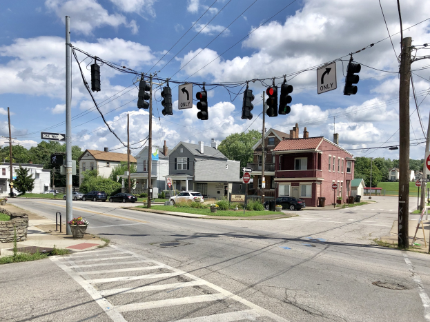 Stadtstraße mit Zebrastreifen, Ampeln, Fahrzeugen, Strommasten, Schildern, blühenden Pflanzen, Gras, Bäumen, Gebäuden mit Fenstern und einem bewölkten Himmel.