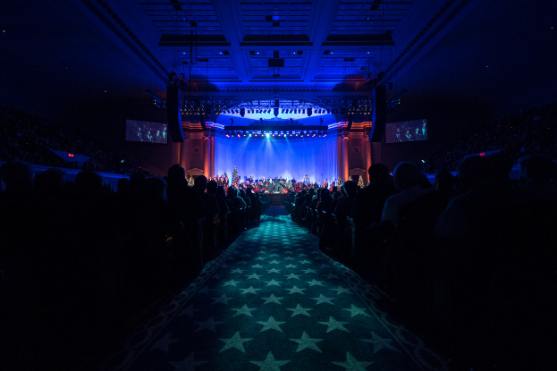 Ein großer Auditorium mit sitzenden Menschen, eine Bühne mit Musikern, die Instrumente spielen, und eine Decke mit Lichtern und Fernsehern, was einen Konzertsaal für die Nationale Convention 2019 vermuten lässt.