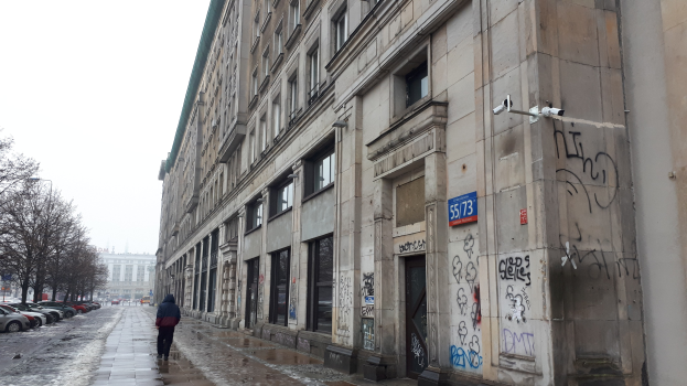 Eine Person, die auf einem Gehweg vor einem Gebäude mit Graffiti an der Wand entlanggeht, mit parkenden Fahrzeugen, Bäumen und Gebäuden im Hintergrund unter einem Himmel.