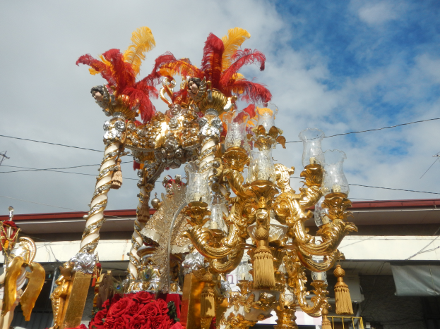 Ein prunkvoller gold-roter Festwagen mit Blumen und Dekorationen bei einem Karnevalsumzug, mit einem Gebäude, Strommasten mit Drähten und einem bewölkten Himmel im Hintergrund.