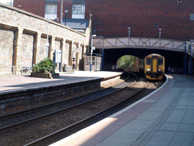 Ein Zug fährt auf den Schienen mit Bahnsteigen auf beiden Seiten, einer Hauswand links, Straßenlaternen, Pflanzen, Bänken, einer Kabine, einer Brücke über die Bahnsteige und Gebäuden und Masten im Hintergrund.