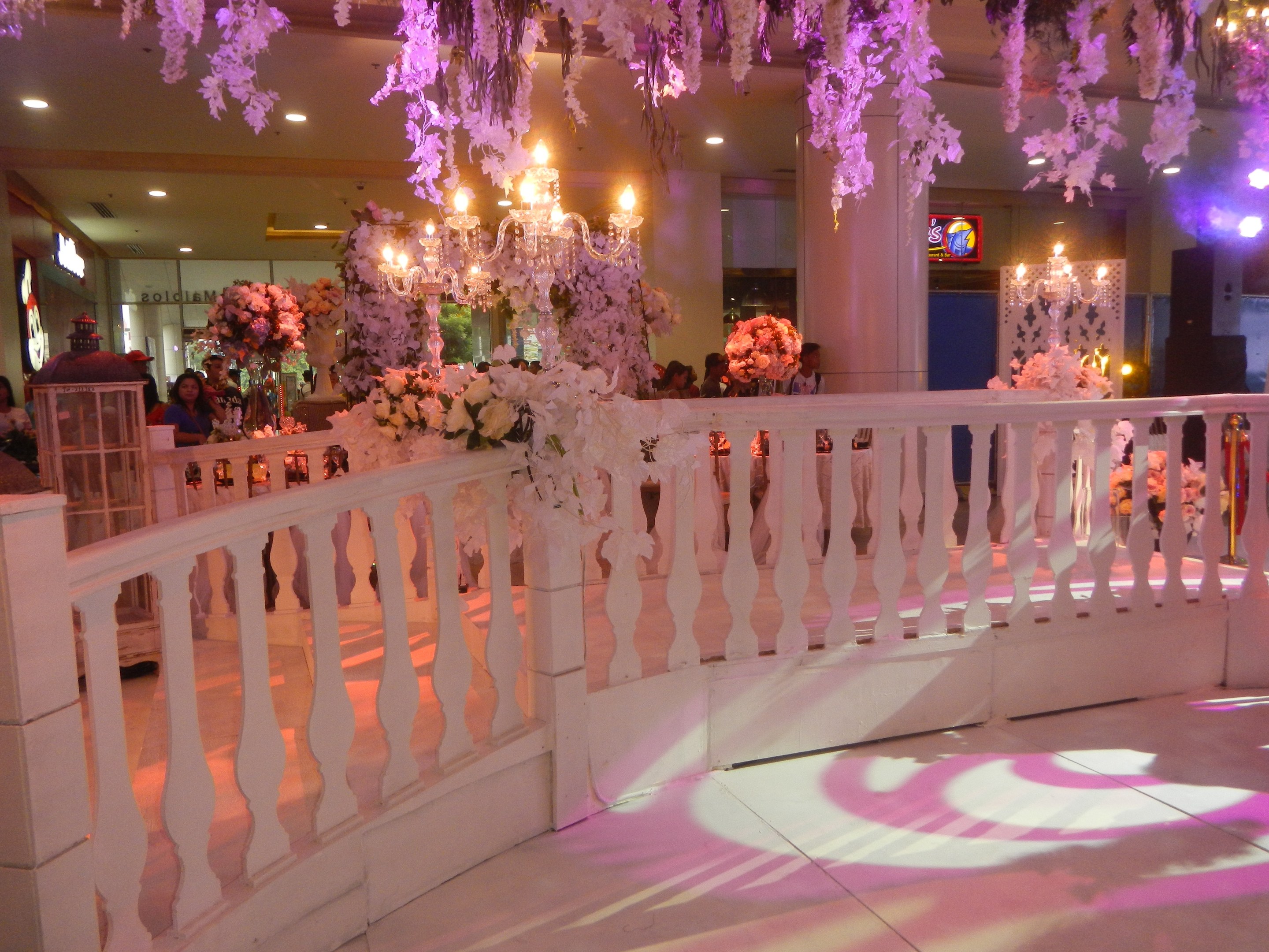 Weiße Reling mit floralen Dekorationen und hängenden Kronleuchtern bei einer Hochzeitsfeier, Menschen versammelt, beleuchtete Säulen im Hintergrund und ein Bildschirm auf der rechten Seite.