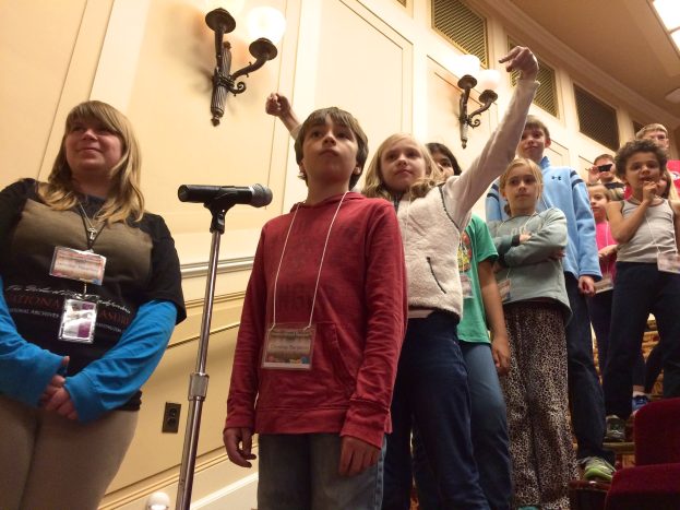 Gruppe von Kindern, die vor einem Mikrofon singen, mit einem Erwachsenen daneben, Stühlen auf der rechten Seite und beleuchteter Wand im Hintergrund.