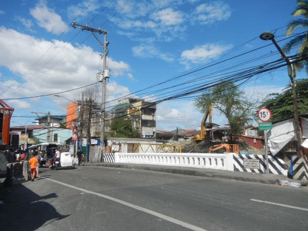 Eine Stadtstraßenszene mit Fahrzeugen, Fußgängern, Strommasten, Schildern, Bäumen, Gebäuden und einer Baustelle unter einem bewölkten Himmel.