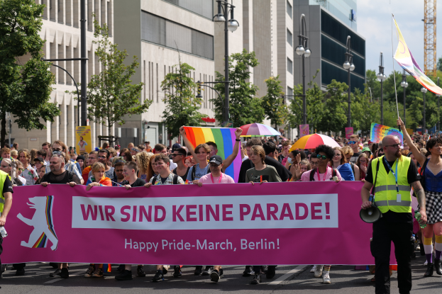 Eine Gruppe von Menschen, die eine rosa Fahne mit der Aufschrift "Happy Pride March Berlin" tragen, geht eine Straße entlang, einige halten Schirme und andere Flaggen, vor dem Hintergrund von Laternenpfählen, Bäumen, Gebäuden mit Fenstern, einem Turm und einem klaren blauen Himmel.