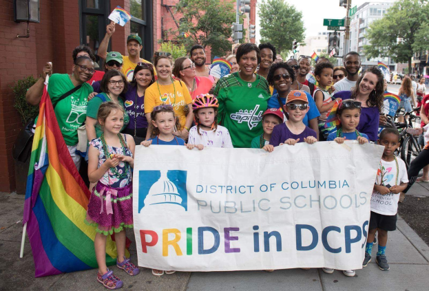 Gruppe von Menschen, die draußen stehen und ein Banner halten, auf dem "District of Columbia Public Schools Pride in DCPS" steht, mit Kindern, die eine Fahne halten, einem Radfahrer, Bäumen, Gebäuden, Straßenschildern, Ampeln und einem bewölkten Himmel.