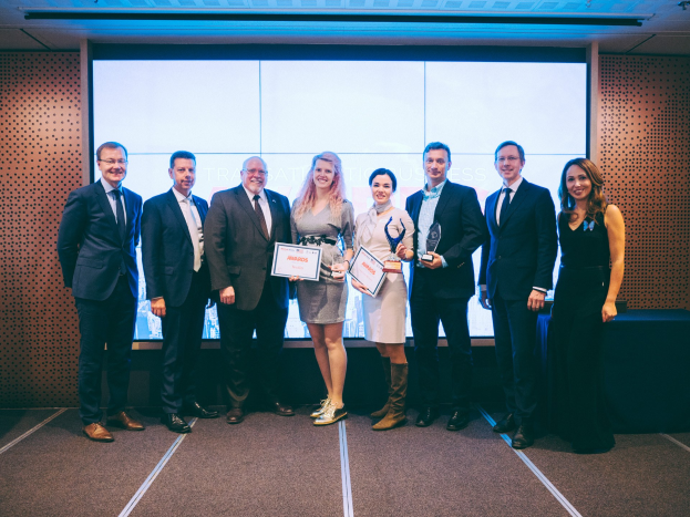 Gruppe von lachenden Menschen mit Urkunden vor einem Bildschirm, mit einem schwarzen bedeckten Tisch und einem wandmontierten Objekt im Hintergrund, Gewinner der International Business Awards 2018.