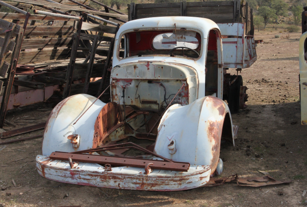 Ein alter, rostiger Lastwagen in einem schlechten Zustand steht im Dreck, umgeben von einem Holzzaun und Bäumen im Hintergrund.