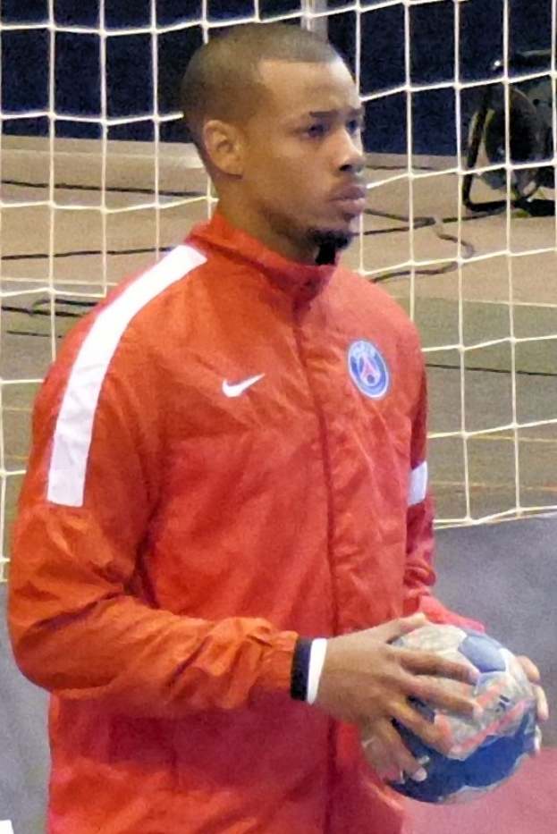 Ein Mann in einem blauen Hemd und Shorts steht vor einem Tor, hält einen Fußball und hat einen entschlossenen Gesichtsausdruck.