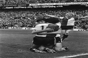 Drei Fußballspieler umarmen sich auf dem Boden und feiern ein Tor, während eine jubelnde Menge und schwenkende Fahnen im Hintergrund zu sehen sind, dargestellt in Schwarz-Weiß.