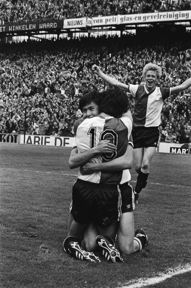 Drei Fußballspieler umarmen sich auf dem Boden und feiern ein Tor, während eine jubelnde Menge und schwenkende Fahnen im Hintergrund zu sehen sind, dargestellt in Schwarz-Weiß.