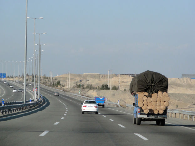 Ein Holzlastwagen mit einer großen Ladung Holz fährt auf einer Autobahn mit Leitplanken, Laternen, Schildern, Bäumen und Sand, mit Hügeln und einem klaren blauen Himmel im Hintergrund.