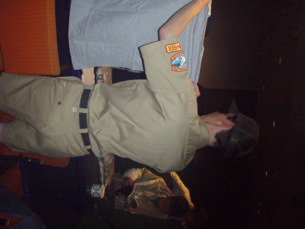 Ein Polizeibeamter hält ein T-Shirt in den Vordergrund, ein Junge hält im Hintergrund ein Buch und einen Becher und steht neben einem Schreibtisch und Lampen.