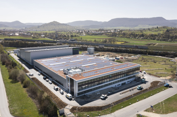 Luftaufnahme eines großen Gebäudes mit Solarmodulen auf dem Dach, umgeben von Fahrzeugen, Bäumen, Gras, Polen und einem klaren blauen Himmel in der Ferne; das Gebäude ist Deutschlands erste solarbetriebene Fabrik.