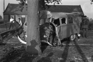 Ein Schwarz-Weiß-Bild eines auf der Straße liegenden Wagens mit einem Baum im Vordergrund, einigen Menschen in der Nähe und Häusern, Bäumen und einem Mast im Hintergrund.