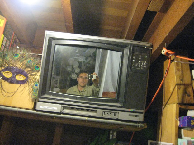 Ein Fernsehbildschirm, der eine Person mit Pfauenfedern, Büchern und Schachteln zeigt.