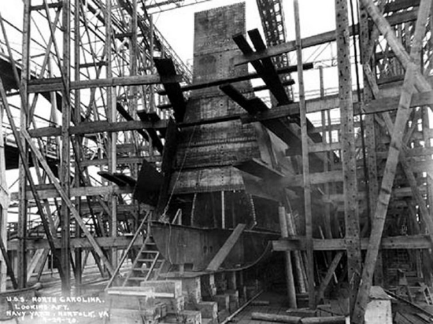 Schwarzes und weißes Foto eines Schiffes in einem Schiffbau, mit hölzernen Pfählen und Treppen im Vordergrund und Himmel im Hintergrund, mit dem Text "Marinebewaffnung" in der linken unteren Ecke.