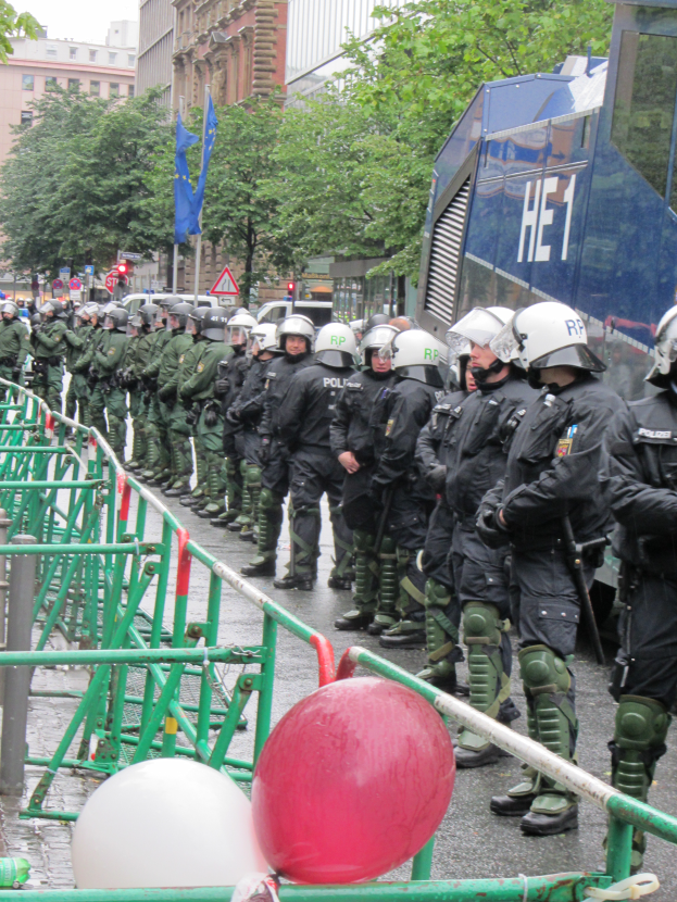 Eine Gruppe von Polizisten in Helmen und mit Waffen vor einer Absperrung auf einer Straße mit Geländern und Luftballons, mit Bäumen, Gebäuden, Fahnen und einem klaren blauen Himmel im Hintergrund.
