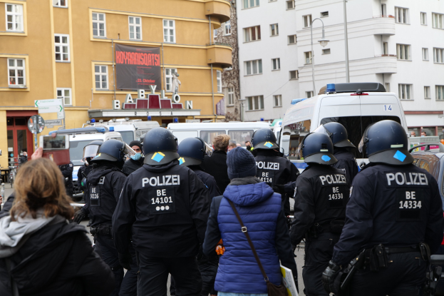 Polizisten in Uniform stehen vor einer Menge mit Helmen und Jacken während einer Demonstration in Berlin, Deutschland, mit Fahrzeugen, Gebäuden, Laternenmasten und einer Fahne im Hintergrund.