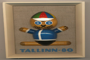 Ein hölzernes Spielzeug mit einer blauen und roten Mütze, beschriftet mit "Tallinn-80", in einem weißen Rahmen.