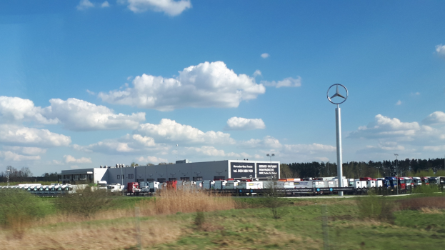 Ein Mercedes-Benz-Werk in Deutschland mit Fahrzeugen auf der Straße, umgeben von Grünflächen, Industriegebäuden und einem Logo-Mast unter einem bewölkten Himmel.