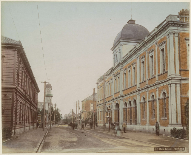 Altes Schwarz-Weiß-Foto einer Stadtstraße mit Gebäuden, Versorgungsmasten, Fahrzeugen, Passanten und Bäumen unter einem Himmel, mit Text unten.