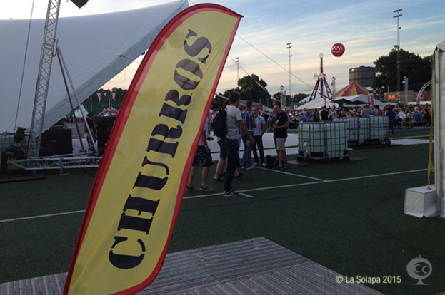 Eine Karnevals Szenerie mit Zelten, einem zentralen Mast, Menschen, Bäumen, Strommasten, Straßenbahnen auf der Straße und einem Eisenständer auf der linken Seite vor einem Himmel-Hintergrund.