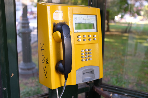 Ein gelbes Münztelefon auf einem Metallpfahl in der Vorderansicht, mit einer unscharfen Sicht auf einen Pfahl, Bäume und Gras durch ein Glasfenster im Hintergrund.