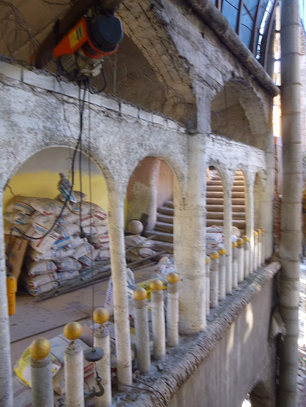 Altes Gebäude unter Renovierung mit architektonischen Bogengängen, Pfeilern und Stufen, umgeben von Werkzeugen, Materialien und verstreuten Gegenständen, einschließlich einer Maschine und Drähten oben und Gegenständen auf dem Boden rechts.