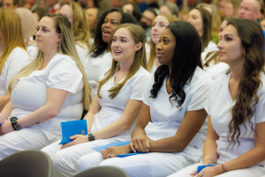 Eine Gruppe von Frauen in weißen Kleidern hält blaue Gegenstände in der Hand und sitzt in einem Hörsaal, der Hintergrund ist unscharf und lenkt die Aufmerksamkeit auf die Frauen, die Teil eines Pflegeprogramms zu sein scheinen.