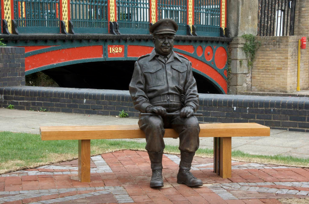 Eine eiserne Statue eines Mannes sitzt auf einer Kartonbank, hinter ihm eine brückenartige Konstruktion mit der goldenen Jahreszahl 1829, flankiert von schwarzen Backsteinwänden und Gittern.