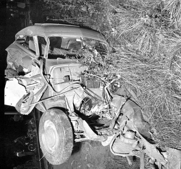 Schwarzes und weißes Foto eines schwer beschädigten Autos mit eingedrückter Front und zerbrochenen Fenstern am Straßenrand, mit einem Baum und verstreuten Gegenständen im Hintergrund.