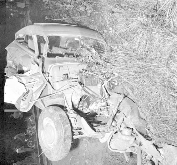 Schwarzes und weißes Foto eines schwer beschädigten Autos mit eingedrückter Front und zerbrochenen Fenstern am Straßenrand, mit einem Baum und verstreuten Gegenständen im Hintergrund.