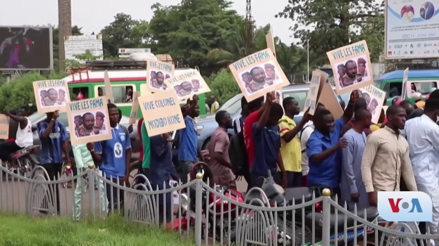 Eine Gruppe von Menschen mit Schildern steht vor einem Zaun, umgeben von Fahrzeugen, B├Ąumen, Polen, Plakaten und einem Bildschirm, mit Gras am Boden und dem Himmel im Hintergrund, scheinbar protestieren sie gegen eine Regierungsentscheidung, die Video-Konferenzen verbietet.