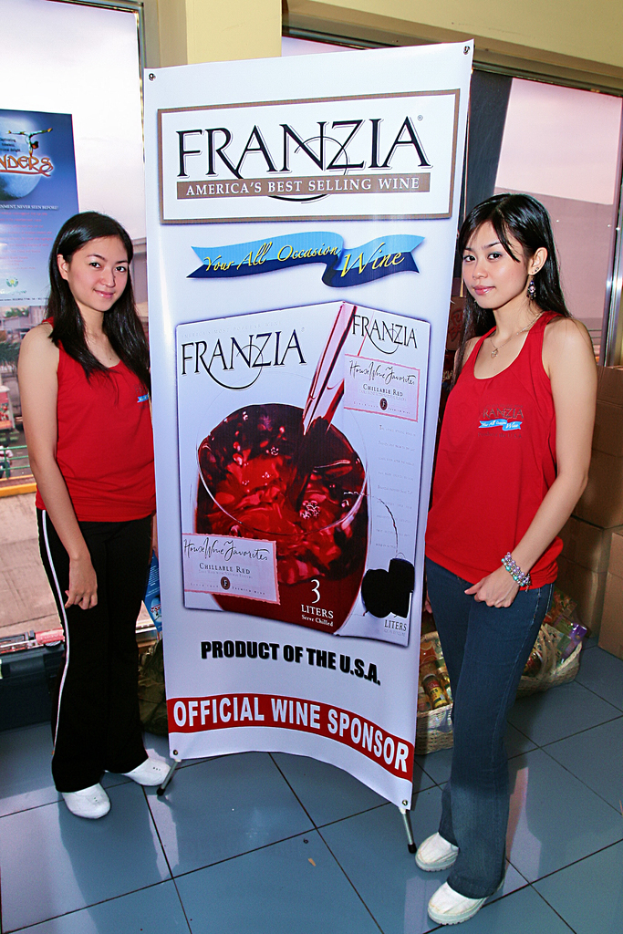 Zwei Frauen neben einem Plakat für Franzia, Americas best-selling wine, mit einem Glasfenster im Hintergrund.