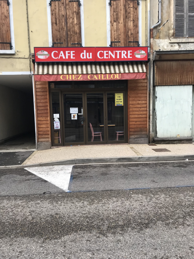 Ein zweistöckiges Gebäude mit einer 'Cafe du Centre'-Aufschrift an der Seite, das Fenster, eine Tür und eine Straße vorne mit einer kleinen Tafel zeigt.