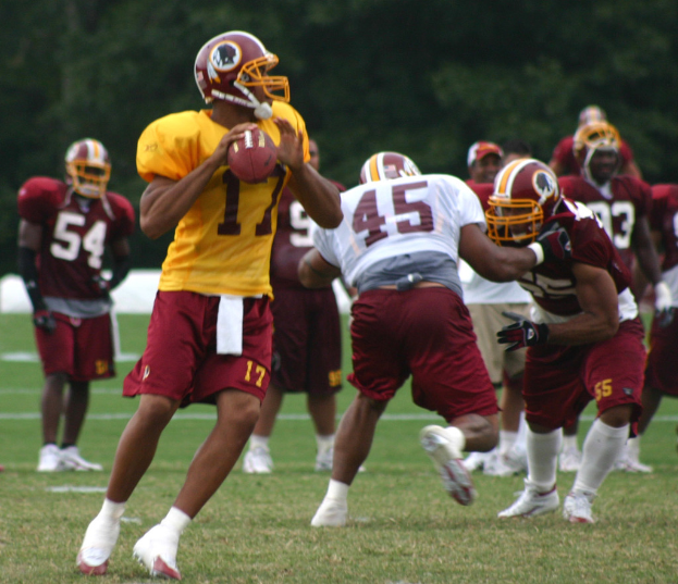 Eine Gruppe von Menschen in Trikots, Helmen und weißen Schuhen spielt American Football auf einem grasbewachsenen Feld mit Bäumen im Hintergrund.