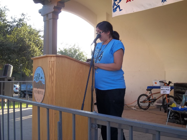 Eine Frau in einem blauen T-Shirt steht vor einem Mikrofon, mit Bäumen, Himmel, Autos, einem Banner und einem kleinen Fahrrad im Hintergrund.