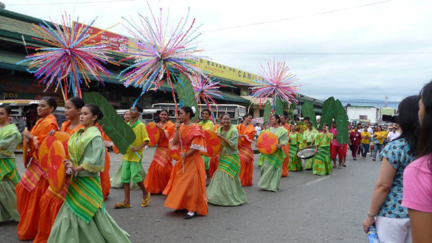 Eine Gruppe von Menschen in bunten Kostümen marschiert auf einer Straße während eines Karnevalsumzugs, mit einer Frau auf der rechten Seite, die eine Flasche hält, und Fahrzeugen, Gebäuden, Bäumen, Polen, Drähten und Himmel im Hintergrund.