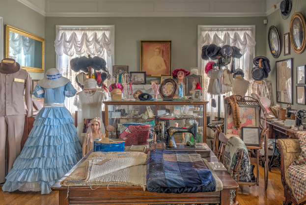 Ein Raum im Museum of Fine Arts in St. Louis, Missouri, gefüllt mit Schaufensterpuppen in verschiedenen Outfits, einem Tisch mit Kleidung und einer Nähmaschine, Stühlen mit Kissen, einem Schrank mit Gegenständen, Fotorahmen an den Wänden, Fenstern mit Vorhängen und einer Decke.