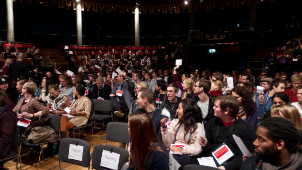 Eine Gruppe von Menschen, die auf Stühlen sitzen und Papiere halten, tragen ID-Karten und sind von Säulen, Lampen, Lautsprechern und zusätzlichen Stühlen umgeben.
