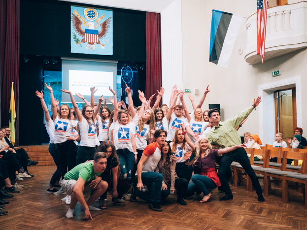 Eine Gruppe von Menschen, die vor einer sitzenden und stehenden Menge posiert, mit einem Bildschirm, Vorhängen, Fahnen, einem Schild, einer Tür, einem Geländer und einer Decke mit Deckenleuchten im Hintergrund.
