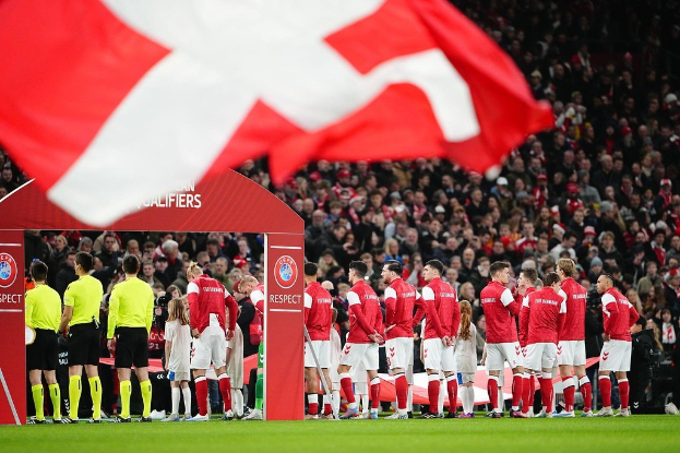 Eine Gruppe von Menschen auf einem Fußballfeld mit einer roten und weißen Flagge im Vordergrund, einem Bogen mit der Aufschrift "Bayern München vs. Bayern München - Wett-Tipps & Vorschau" im Hintergrund und einer großen Menge im Stadion.