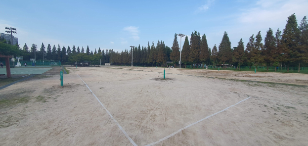 Ein Beachvolleyballplatz in einem Park, umgeben von Pfählen, einem Zaun, Bäumen, Gebäuden, Straßenlaternen.