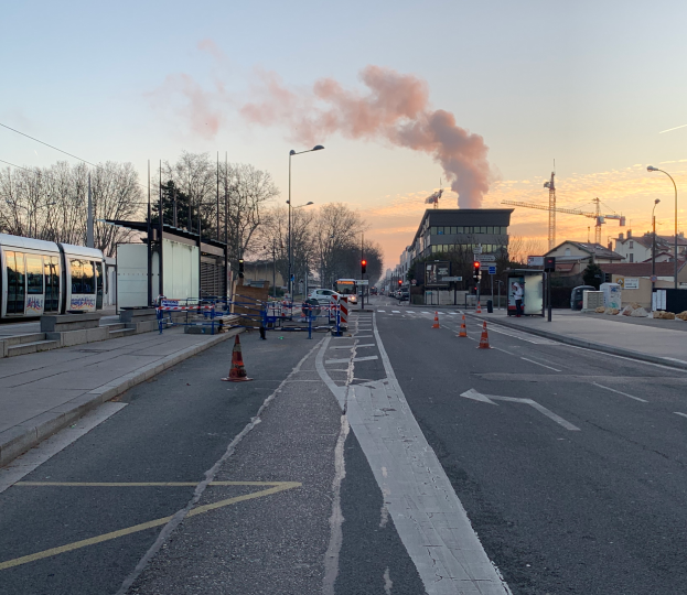 Eine Stadtstraße mit Verkehrskegeln, einem Bus, Laternenpfählen, Bäumen, Gebäuden, Kränen und Rauch, der aus einem entfernten Gebäude aufsteigt, unter einem klaren Himmel.