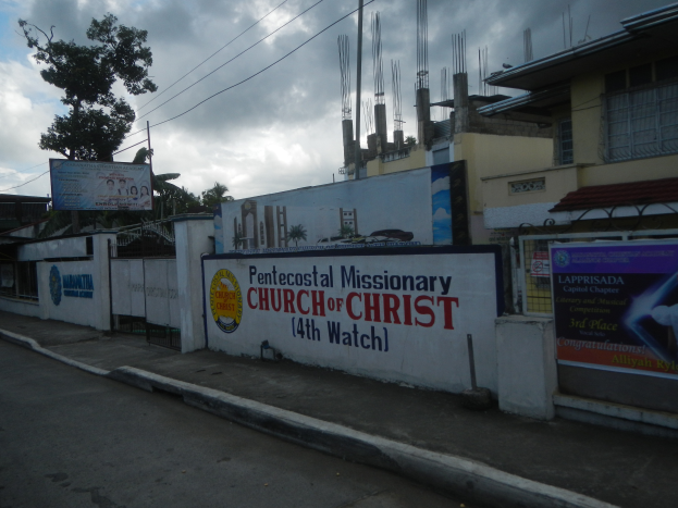 Die Pfingstmissionarische Kirche Christi 14. Wache steht mit einer Straße im Vordergrund, einer Wand mit Bannern und Text, Gebäuden, Bäumen, Polen, Drähten und einem bewölkten Himmel im Hintergrund.