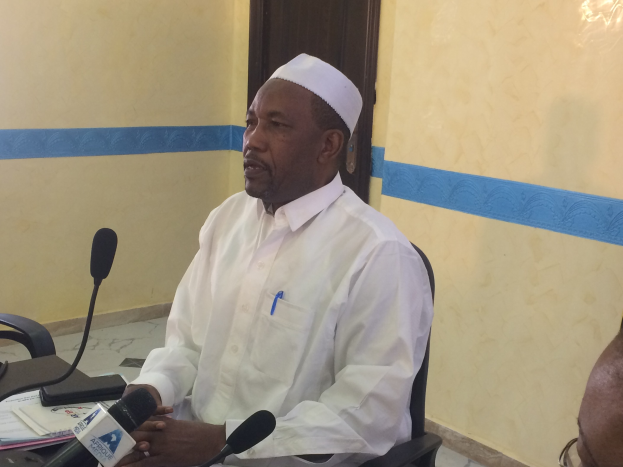 Ein Mann in weißem Anzug und Hut sitzt an einem Tisch mit Mikrofonen, spricht, während Papiere und Gegenstände auf dem Tisch liegen, mit einer anderen Person mit Brille rechts von ihm in einem Raum mit einer Wand und einer Tür im Hintergrund.