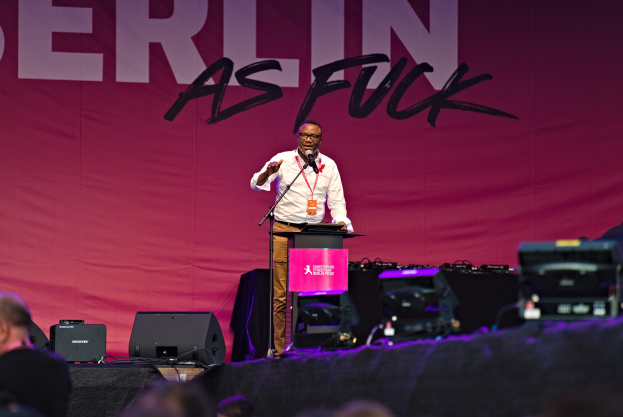 Mann in weißem Hemd und braunen Hosen hält Mikrofon und spricht zu einer Menge, hinter ihm ein pinkes Banner mit der Aufschrift 'Berlin as Fuck' und Lautsprecher auf der Bühne.