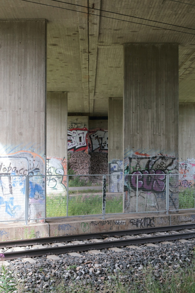 Eisenbahnschiene mit Graffiti an einer Seitenstruktur, umgeben von Steinen, Gras, einem Zaun, Pfeilern, Oberleitungen und einem Dach.